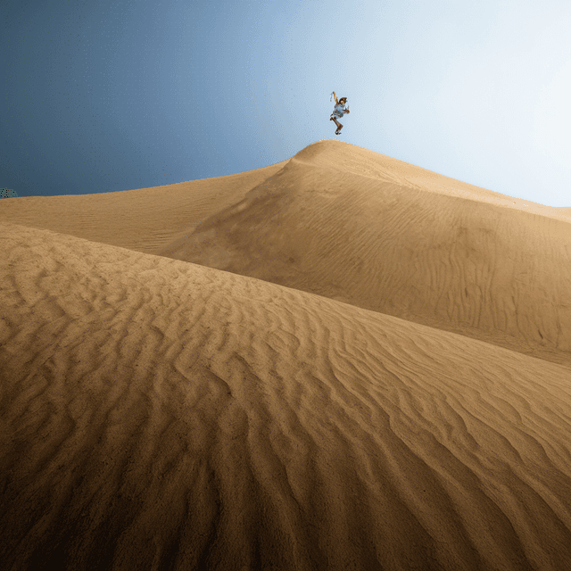 The image captures a dynamic scene of a person leaping off the crest of a large sand dune in an expansive desert landscape.