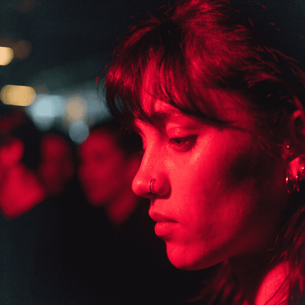 A very tight close-up shows a young woman’s face in profile turned slightly downward, filling the right half of the frame against a nearly black, indistinct background.