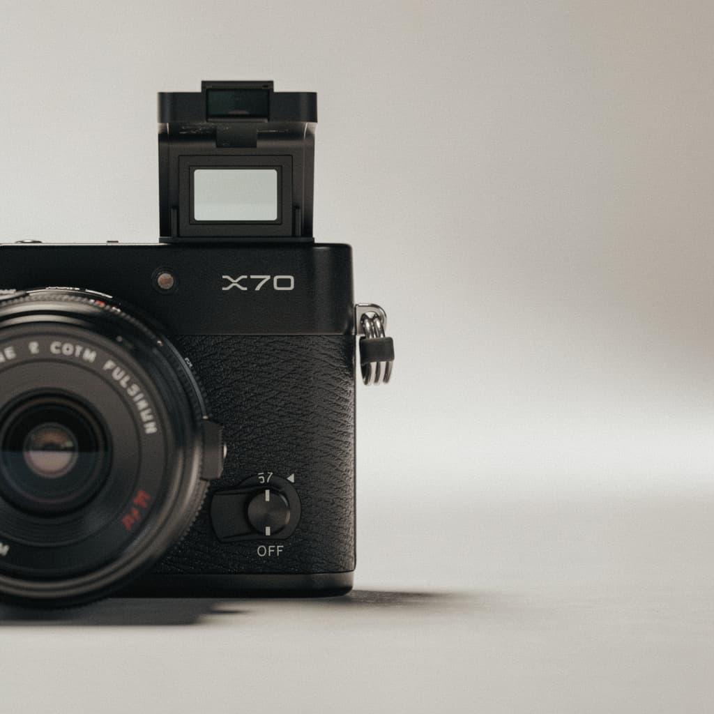 A compact black Fujifilm X70 camera sits close to the viewer on a plain, lightly textured surface, framed from a slightly elevated front-left angle so the lens and top plate dominate the lower half of the composition.
