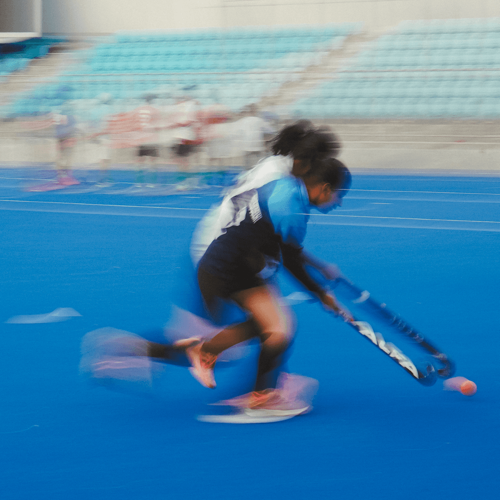 Two field hockey players are captured in a fast, motion-blurred close view on a vivid blue synthetic turf pitch, leaning low as they chase a pink ball near the lower right edge.