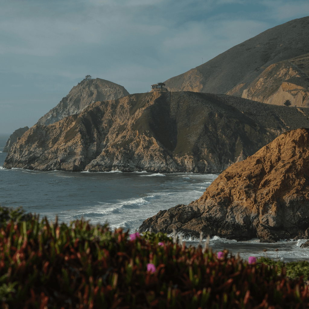 A rugged coastal landscape fills the frame, viewed from a slightly elevated distance across a cove with dark blue-green water and white surf curling along the rocky shore.