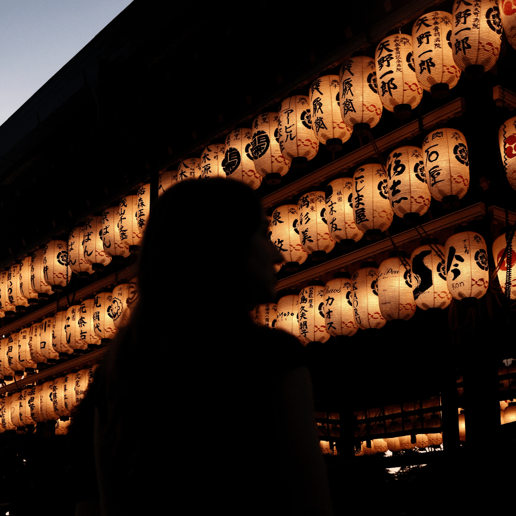A dark exterior setting at dusk or night shows the underside of a traditional building roof at the top left against a small wedge of pale sky, while three tightly packed rows of warm amber paper lanterns fill the upper and right side of the frame.