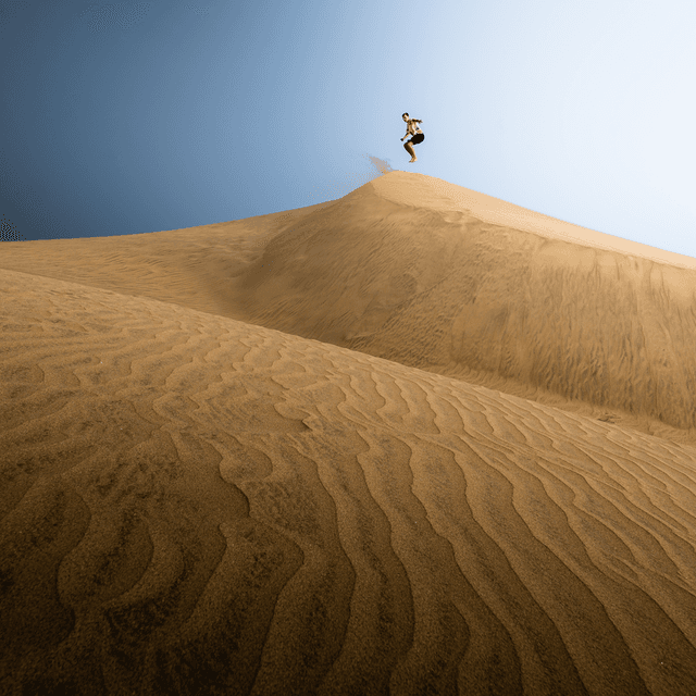 The image captures a dynamic scene of a person leaping off the crest of a large sand dune in an expansive desert landscape.