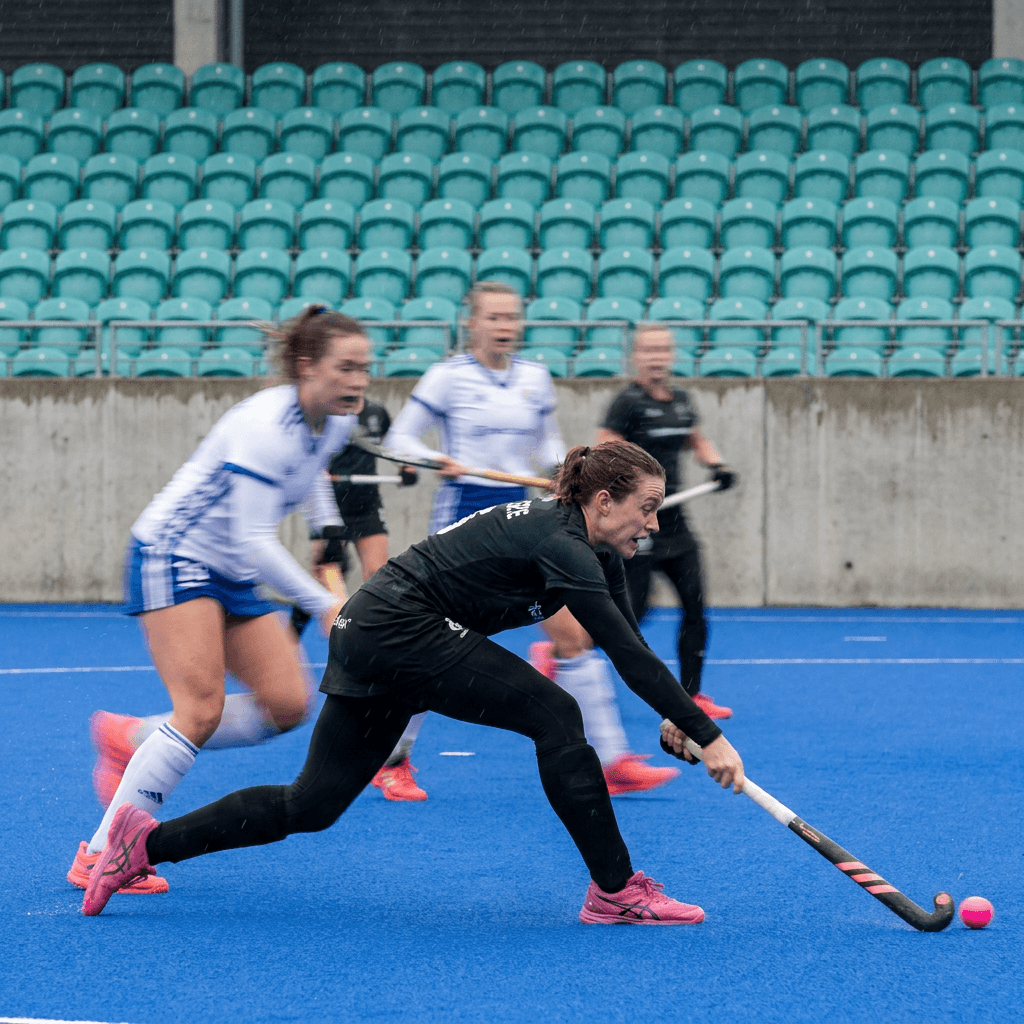Two field hockey players are captured in a fast, motion-blurred close view on a vivid blue synthetic turf pitch, leaning low as they chase a pink ball near the lower right edge.
